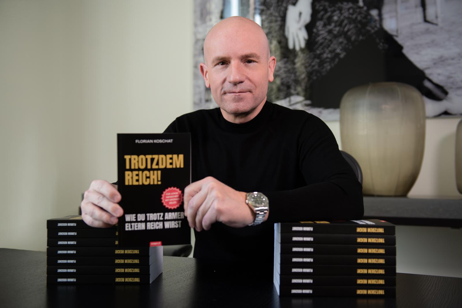 Dr. Florian Koschat hält „Trotzdem reich!“ zwischen zwei Bücherstapeln