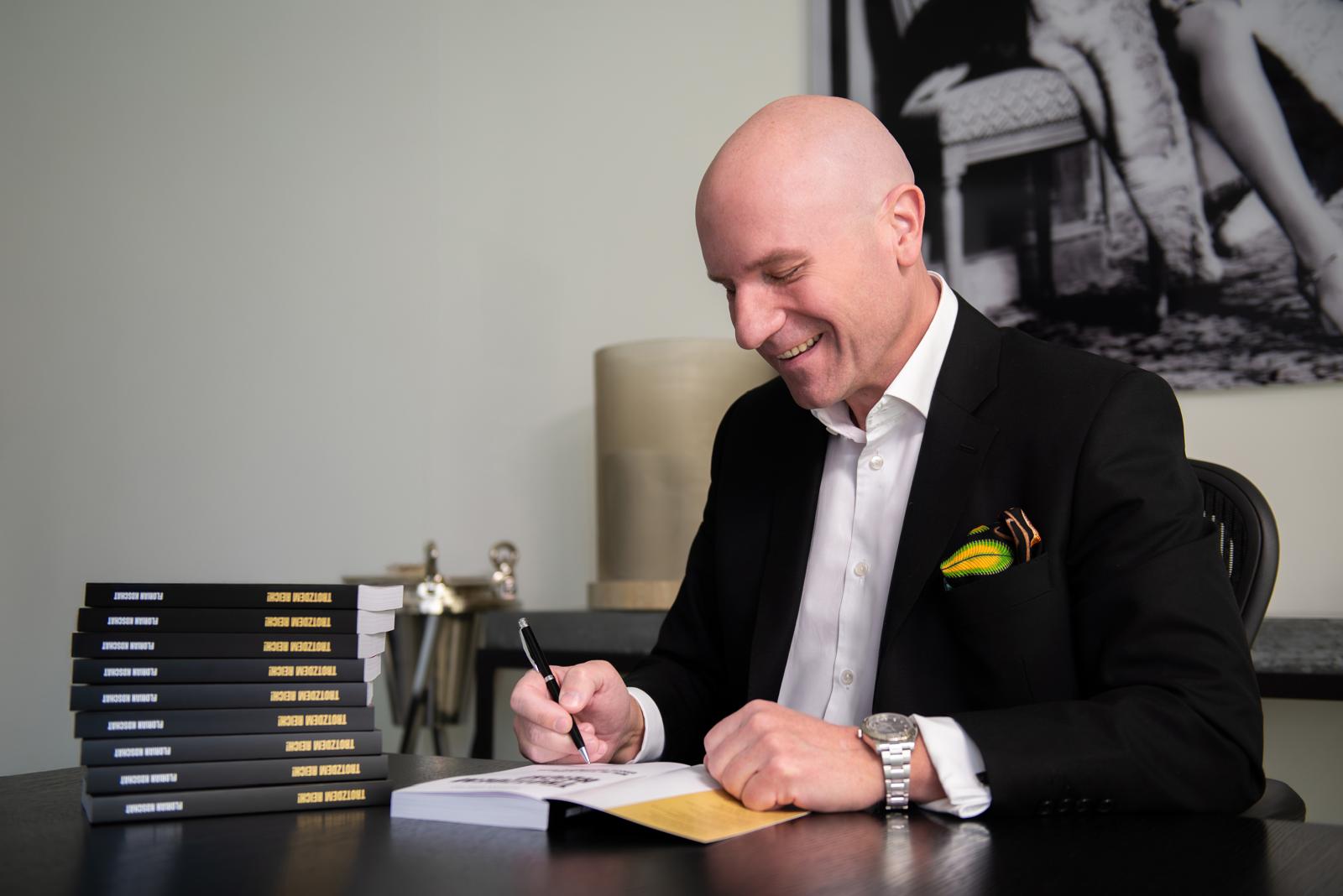 Dr. Florian Koschat signiert lachend sein Buch „Trotzdem reich!“, daneben ein Stapel Bücher